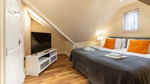 Quarto com televisão em móvel, cama com almofadas laranja, janela com persianas e porta branca.