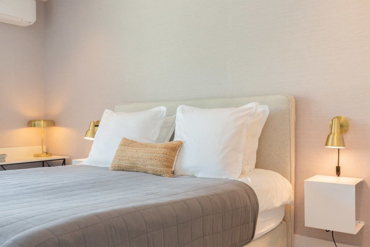 Una cama bien tendida con colcha gris, almohadas blancas y un cojín decorativo color beige. Hay dos mesitas de noche con lámparas, una a cada lado de la cama.