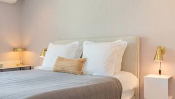 Una cama bien tendida con colcha gris, almohadas blancas y un cojín decorativo color beige. Hay dos mesitas de noche con lámparas, una a cada lado de la cama.