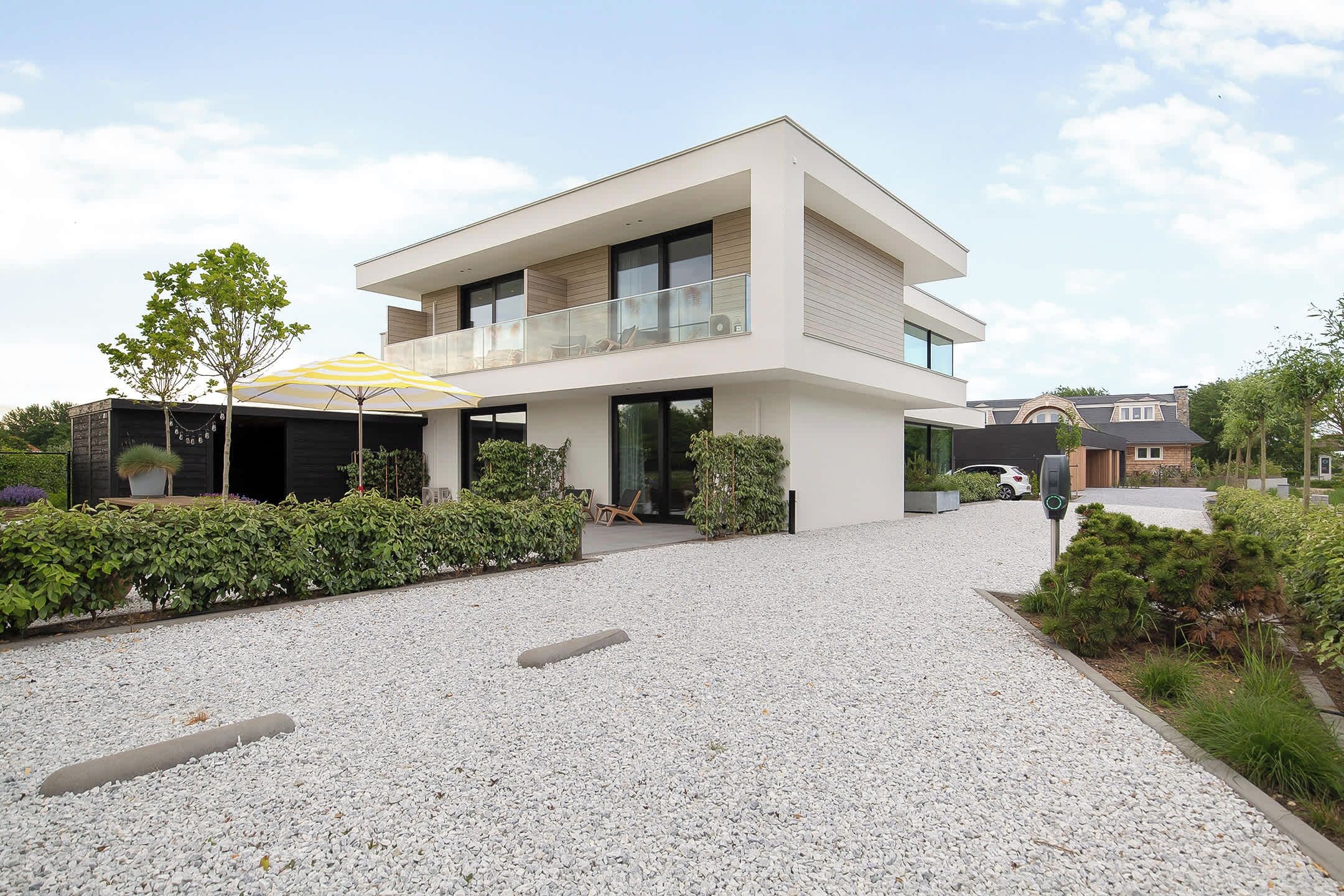 Een modern huis met twee verdiepingen, een overdekte patio, een grindpad en omliggend groen.