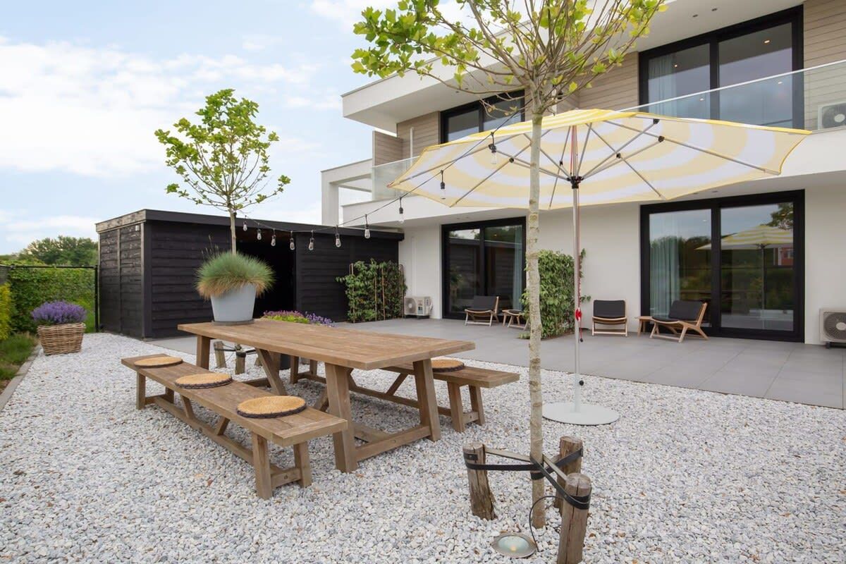 Un patio exterior moderno con una mesa y bancos de madera, rodeado de grava, y una sombrilla grande que proporciona sombra.
