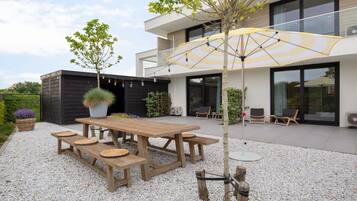 Un patio exterior moderno con una mesa y bancos de madera, rodeado de grava, y una sombrilla grande que proporciona sombra.