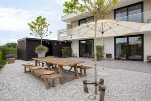 A modern outdoor patio with a wooden table and benches, surrounded by gravel, and a large umbrella providing shade. - Studio near beach in Domburg | Summum Suites 2 (Domburg)