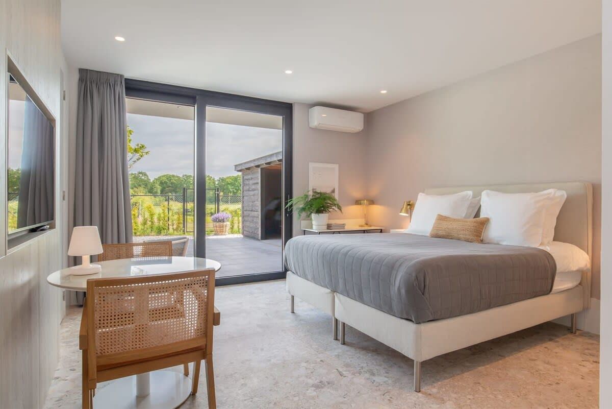 Un dormitorio moderno con una cama grande, una mesita pequeña y una silla. Hay un balcón con vistas a zonas verdes.