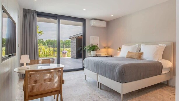 A modern bedroom with a large bed, a small table, and a chair. There is a balcony with a view of greenery. - Studio near beach in Domburg | Summum Suites 2 (Domburg)