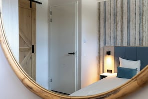 Une chambre à coucher avec un lit, une table de chevet et une lampe fixée au mur. On aperçoit une porte au fond.