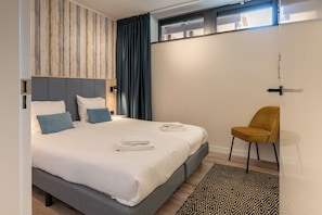 Une chambre à coucher moderne avec un grand lit, une tête de lit dotée de lattes en bois verticales, un rideau foncé, une petite chaise jaune et un tapis à motifs.