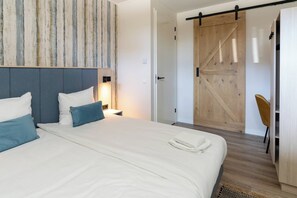 A modern bedroom with a large bed, a wooden sliding door, a chair, and a nightstand with a lamp. - Apartment with sauna near the beach | Duno Lodge 15-A (Oostkapelle)