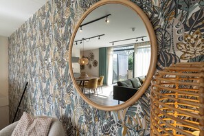 Ein Zimmer mit ornamentalem Wandpapier, einem runden Spiegel, einem Korbstuhl und einem Essbereich mit Stühlen und Tisch.