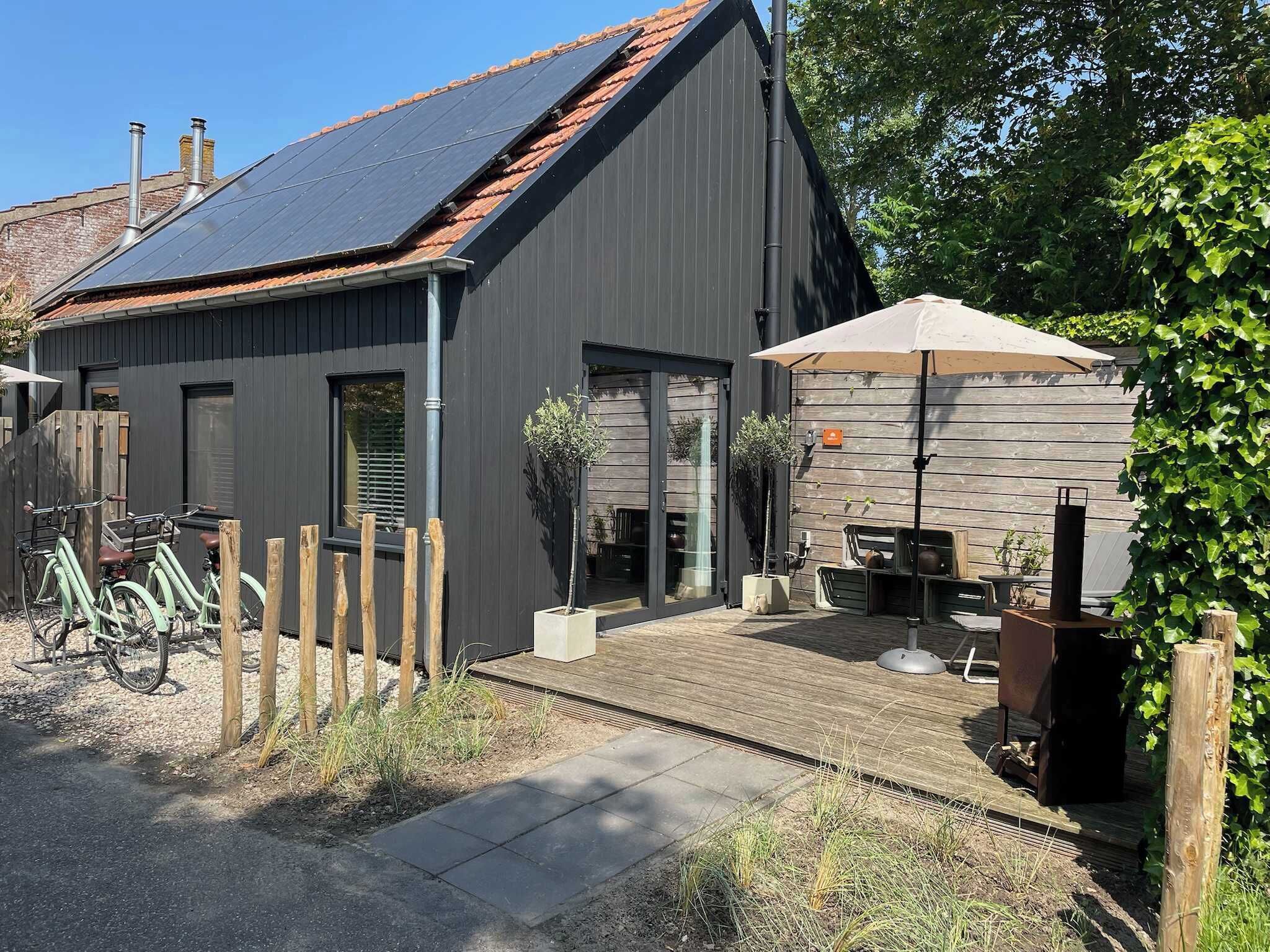 Ein modernes Gebäude mit dunkler Fassade, einem geneigten Dach mit Solarpanels, einer Holzterrasse und einer Patio mit Gartenmöbeln.