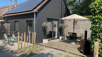 Ein modernes Gebäude mit dunkler Fassade, einem geneigten Dach mit Solarpanels, einer Holzterrasse und einer Patio mit Gartenmöbeln.