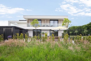Une maison moderne à deux étages, dotée d’un balcon et entourée d’herbes hautes et d’arbres.