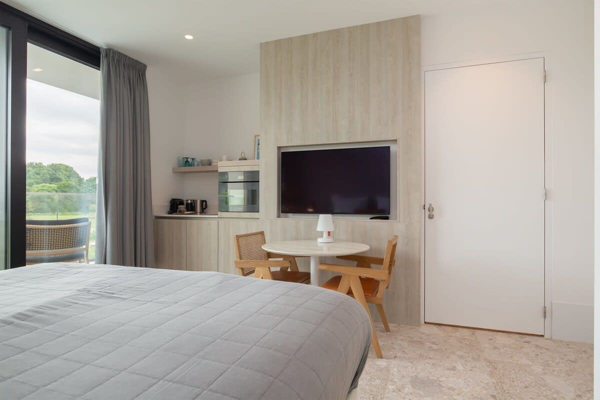 Una habitación de hotel moderna con una cama grande, un área de comedor con mesa y sillas, un televisor montado en la pared y vistas al exterior.