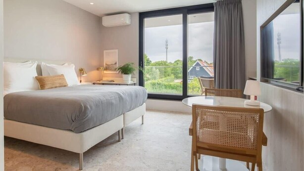 A modern bedroom with a large bed, a dining table, and a view of greenery and a house outside the window.