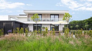 A modern two-story house with a balcony, surrounded by tall grass and trees.