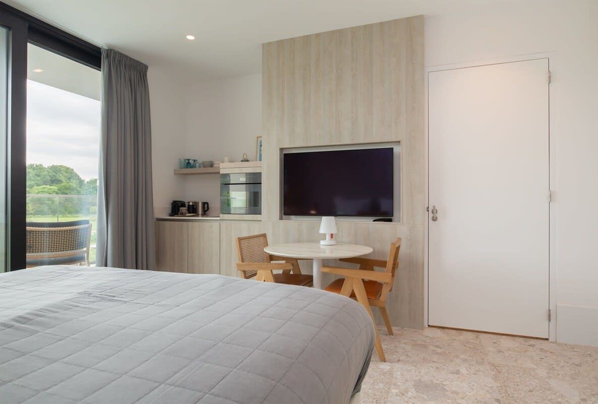 Een moderne hotelkamer met een groot bed, een zithoek met tafel en stoelen, een aan de muur bevestigde televisie en uitzicht op het omliggende landschap.