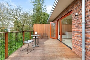 Un balcon doté d’une terrasse en bois, de chaises en métal, d’une table et d’une porte vitrée donnant sur un espace intérieur.