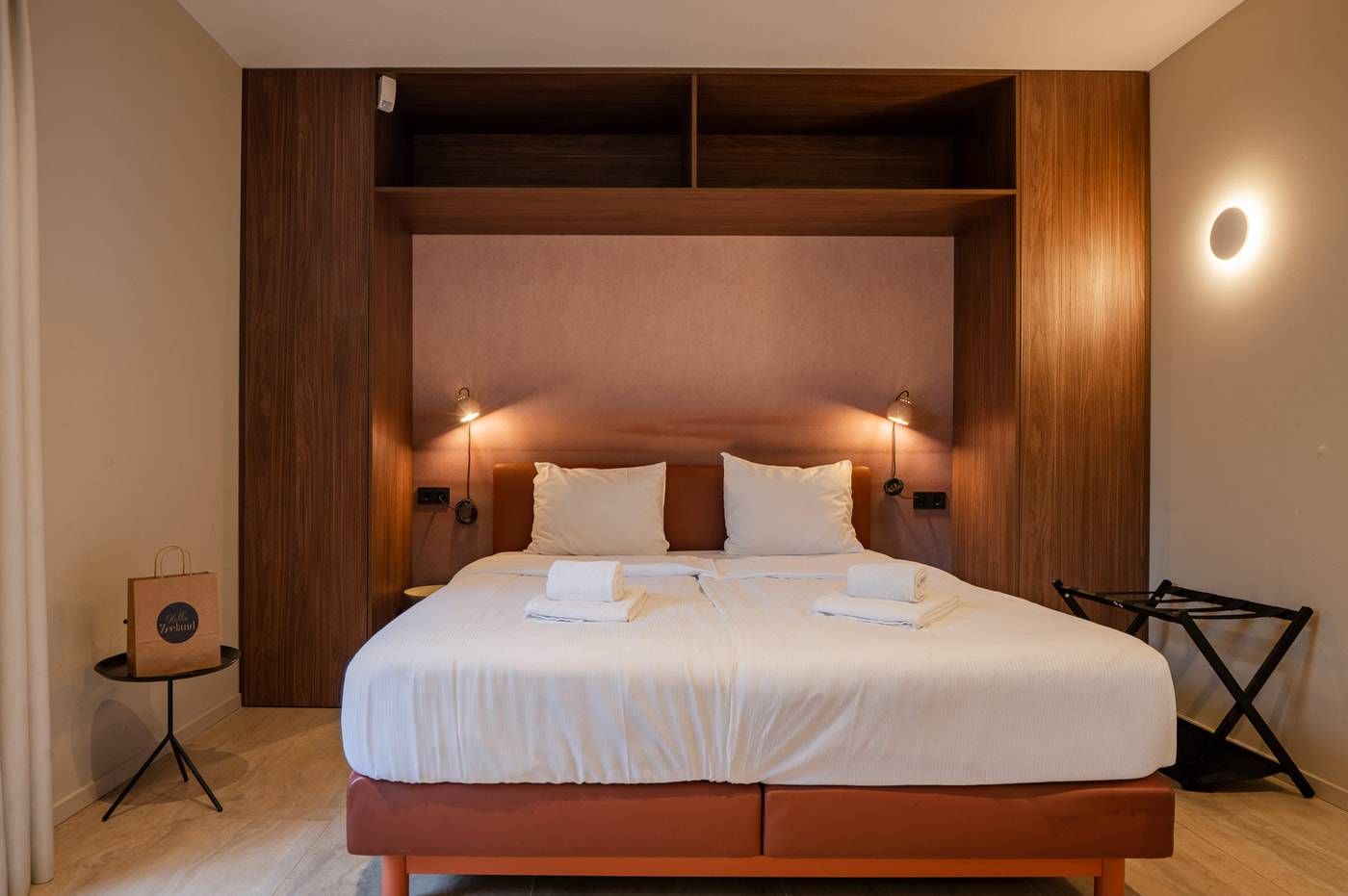 Une chambre d’hôtel avec un grand lit, deux lampes de chevet, une tête de lit en bois et une petite table sur laquelle se trouve un sac.