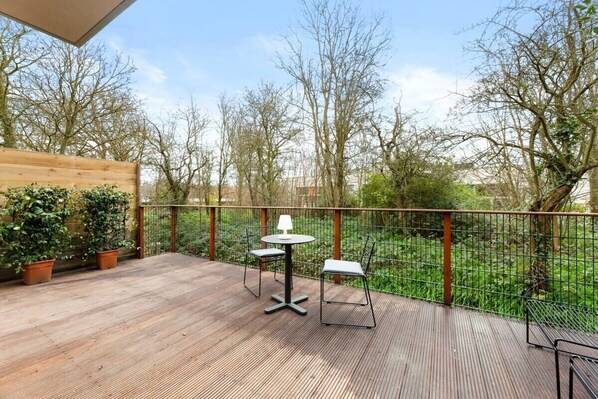 Een terras buiten met een houten vloer, twee tafels met stoelen, en uitzicht op bomen en begroeiing.