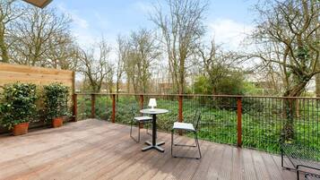 Una terraza al aire libre con piso de madera, dos mesas con sillas y vista a árboles y follaje.