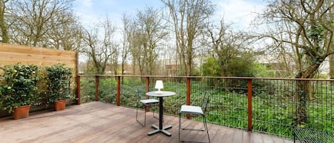Una terraza al aire libre con piso de madera, dos mesas con sillas y vista a árboles y follaje.