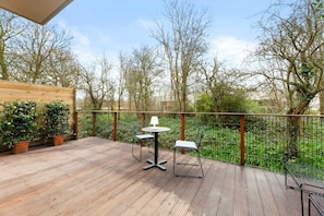 Une terrasse extérieure dotée d’un plancher en bois, de deux tables avec des chaises, et offrant une vue sur les arbres et la verdure.