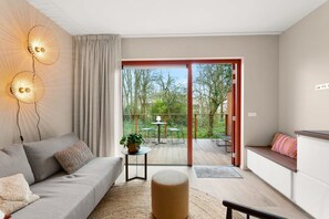 Un salon moderne avec un canapé, un pouf et une petite table sur laquelle est posée une plante. Une porte-fenêtre coulissante donne accès à un patio extérieur aménagé avec une table et des chaises.