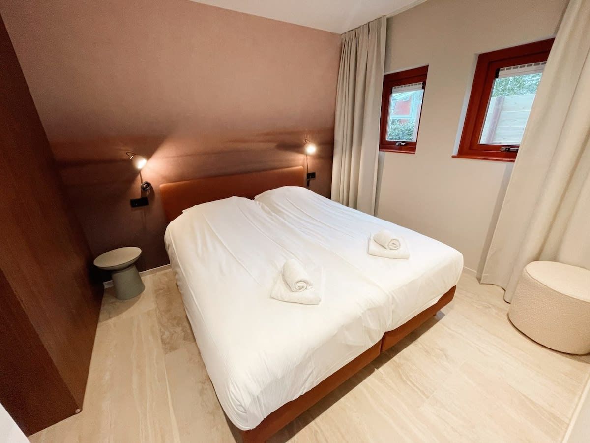 Une chambre d’hôtel avec un grand lit, deux tables de chevet, une fenêtre avec des rideaux et un petit tabouret rond.