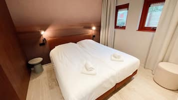 Une chambre d’hôtel avec un grand lit, deux tables de chevet, une fenêtre avec des rideaux et un petit tabouret rond.