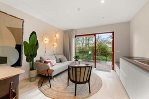 Un salon moderne comprenant un canapé, une table basse et des fauteuils, un coin repas avec une table et des chaises, une cuisine avec un évier et une grande fenêtre donnant sur un espace verdoyant.