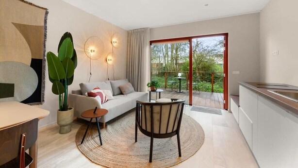 A modern living room with a sofa, coffee table, and chairs, a dining area with a table and chairs, a kitchen with a sink, and a large window with a view of greenery. - Apartment in the centre of Domburg | Duinenburg 10-3 (Domburg)