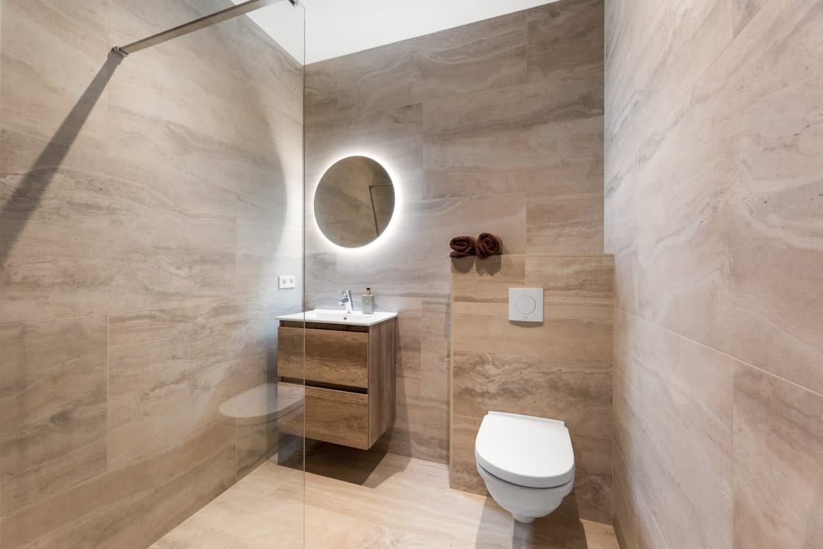 Un baño moderno con cabina de ducha de vidrio, tocador de madera, espejo redondo y retrete.