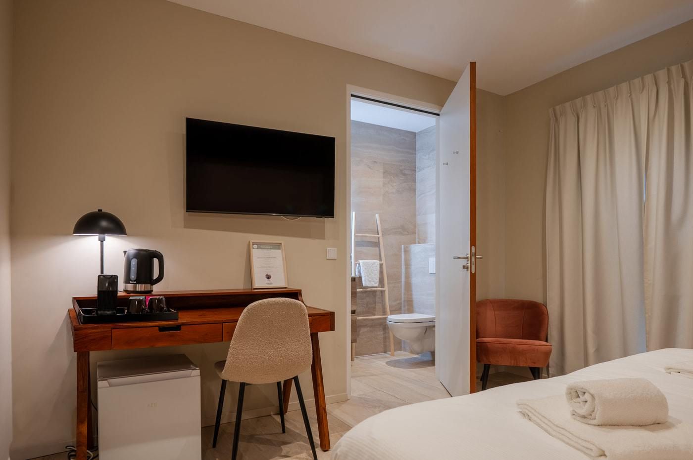 Ein Hotelzimmer mit einem Bett, einem Schreibtisch mit Lampe, einem Fernseher und einem durch eine offene Tür einsehbaren Badezimmer.