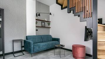 Een moderne woonkamer met een blauwe bank, een klein bijzettafeltje, een rode poef en een trap met houten treden.