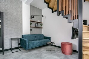 A modern living room with a blue sofa, a small side table, a red ottoman, and a staircase with wooden steps.