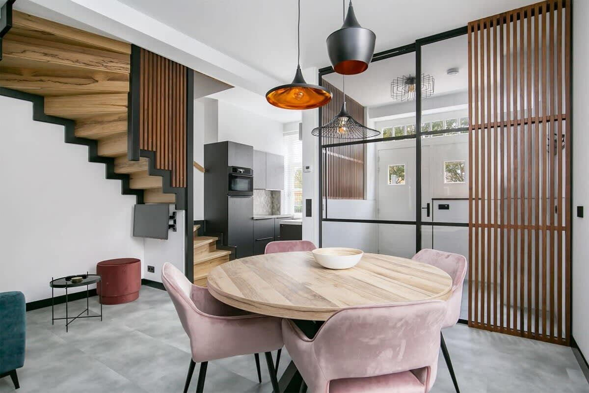 Une cuisine moderne avec un escalier en bois, un coin repas avec une table ronde et des chaises roses, et une porte-fenêtre coulissante donnant sur un balcon.