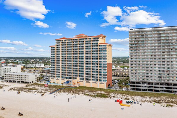 Un hotel moderno de gran altura con múltiples balcones, una playa en primer plano y un cielo azul despejado.