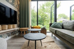 Ein modernes Wohnzimmer mit großem Fernseher, einer Couch, einem runden Tisch und Blick auf Grünfläche.