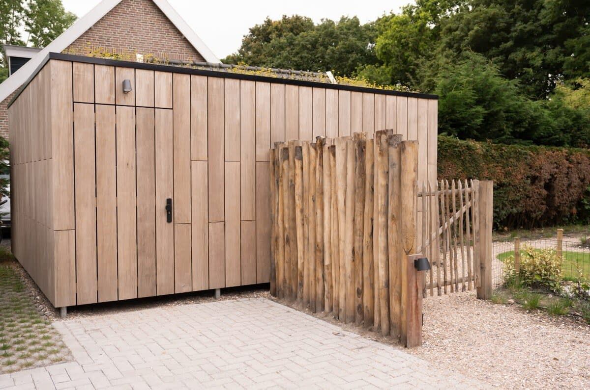 Un abri de jardin en bois, doté d’une porte en métal et d’un portail en bois, situé dans un jardin avec un chemin pavé.
