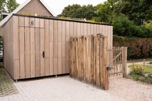 Un abri de jardin en bois, doté d’une porte en métal et d’un portail en bois, situé dans un jardin avec un chemin pavé.