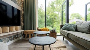 A modern living room with a large TV, a sofa, a round table, and a view of greenery outside. - Apartment near the beach in Oostkapelle | Duno Lodge 15-G (Oostkapelle)