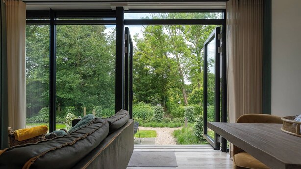 A modern interior with a sofa, dining table, and large glass doors leading to a garden. - Apartment near the beach in Oostkapelle | Duno Lodge 15-G (Oostkapelle)