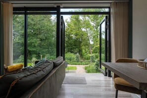 Ein modernes Wohnzimmer mit einer Couch, einem Esstisch und Blick auf einen Garten durch große Fenster.