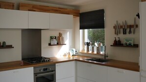 Een moderne keuken met witte kastjes, een houten aanrecht, een gasfornuis, een oven, een spoelbak en een raam met zonwering.