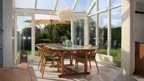 Een oranjerie met een glazen tafel en stoelen, een witte hanglamp en uitzicht op de tuin.