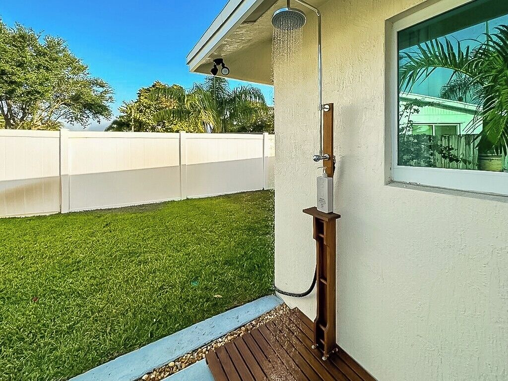 A residential outdoor shower setup with a hose, a wooden deck, and a white fence in the background.
