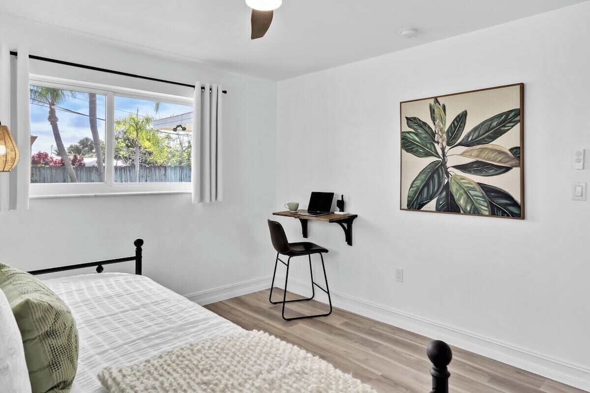 A bedroom with a bed, a desk with a chair, a window with curtains, and a wall-mounted artwork.