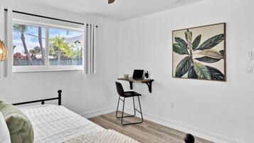 A bedroom with a bed, a desk with a chair, a window with curtains, and a wall-mounted artwork.