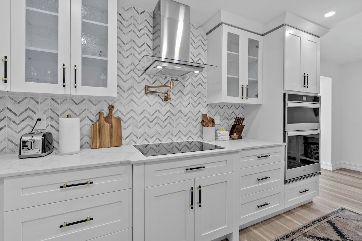 A modern kitchen with white cabinetry, a stainless steel range hood, a built-in oven, and a herringbone-patterned backsplash.
