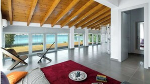 A spacious room with a high ceiling and wooden beams, large windows offering a view of the sea, a red rug, and a chair. - Beachfront villa 100 metres from the sandy beach (Kontokálion)